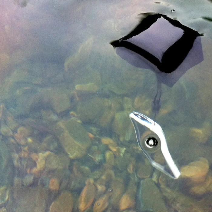 A smartphone with its screen facing up floats in shallow, clear water above rocks, secured by a Catalyst Case US Floating Lanyard - Stealth Black that's attached by a strap and partially above the surface.