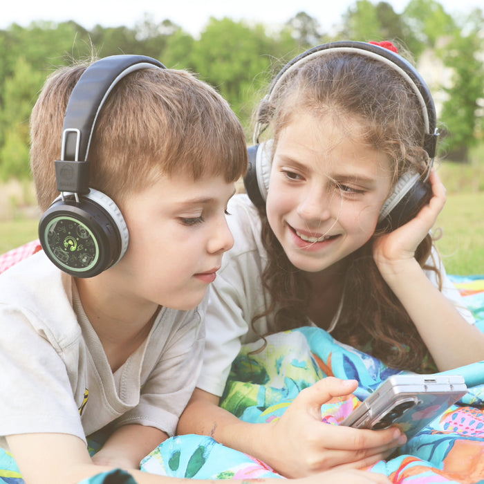 Two children wearing the MEE audio KidJamz Share Bluetooth Headphones on a cell phone while laying at a park