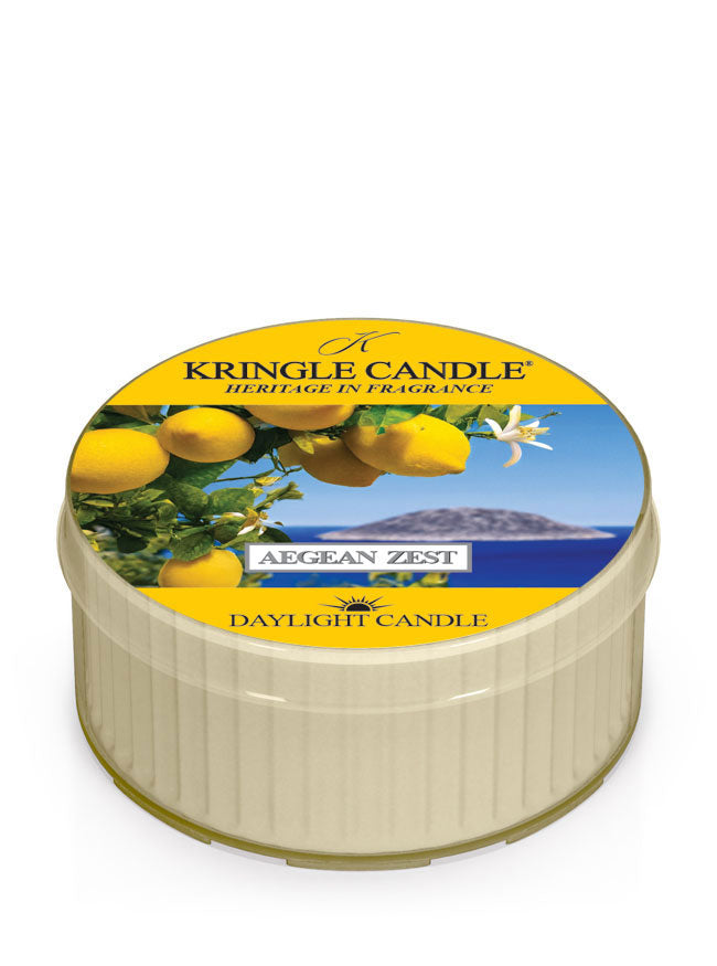 Kringle Candle Aegean Zest Daylight Candle – A small, cream-colored candle in a plastic cup with a vibrant lid design featuring fresh lemons, white blossoms, and a scenic ocean view in the background. The label includes ‘Kringle Candle - Heritage in Fragrance,’ the fragrance name ‘Aegean Zest,’ and ‘Daylight Candle’ branding in yellow and white text.