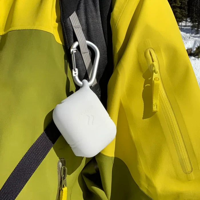 A Catalyst Case US AirPods (Gen 2/1) Waterproof Case with carabiner is clipped to the strap of a yellow and green jacket with a zippered sleeve pocket. Snowy trees can be seen in the background. No further product description is available.