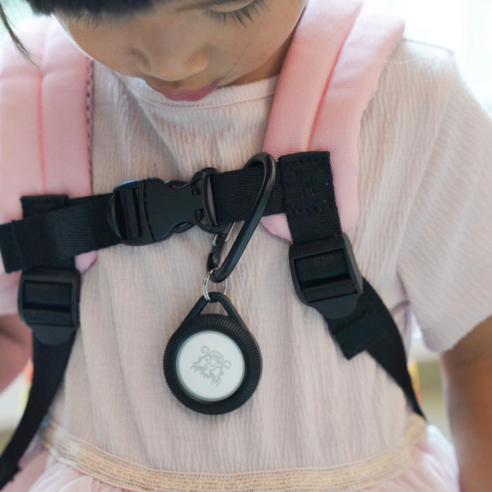 A young child wearing a pale pink dress carries a pink backpack with black straps and a plastic buckle, featuring the Catalyst Case US AirTag - Total Protection Case, Hang-It.