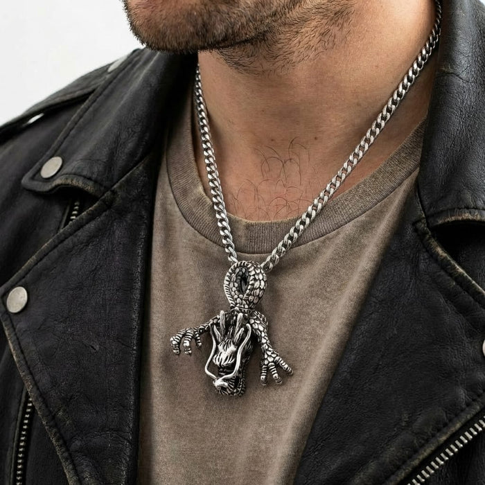 Close-up lifestyle shot of a man wearing a distressed black leather motorcycle jacket and a faded t-shirt, modeling a large, detailed sterling silver dragon head pendant with claws on a silver chain necklace.