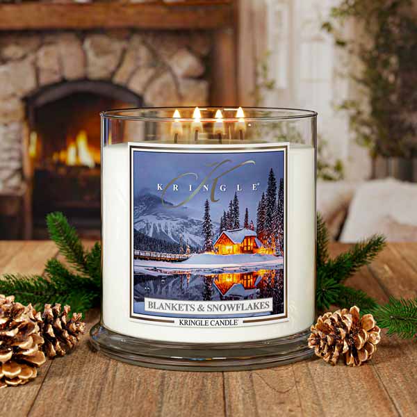 Kringle Candle with a winter scene label on a wooden surface with pinecones and evergreen branches, fireplace in the background.