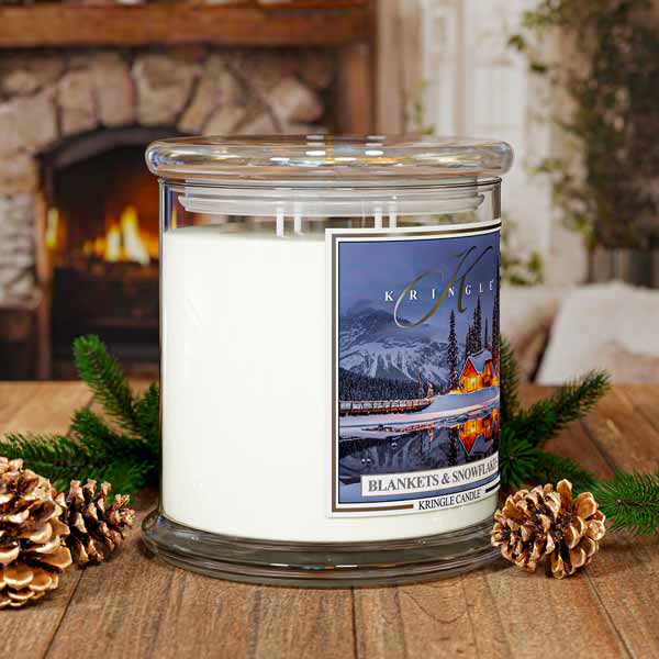 Kringle Candle with a scenic label on a wooden surface with pine cones and evergreen branches, fireplace in the background