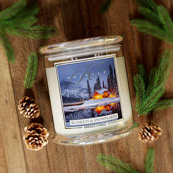 Kringle Candle with winter scene label for Blankets & Snowflakes on a wooden surface with pine cones and branches
