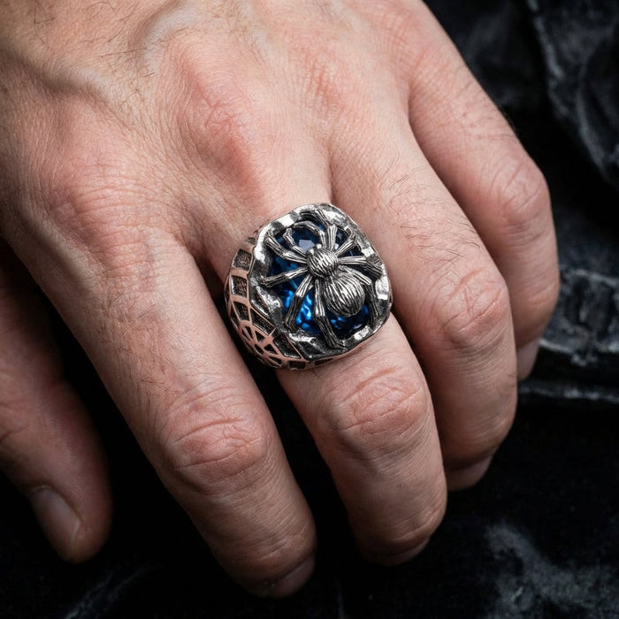 Men's gothic silver spider ring with blue gemstone worn on hand, featuring detailed spider design over a deep blue stone with spiderweb pattern on the band sides, displayed against a dark fabric background