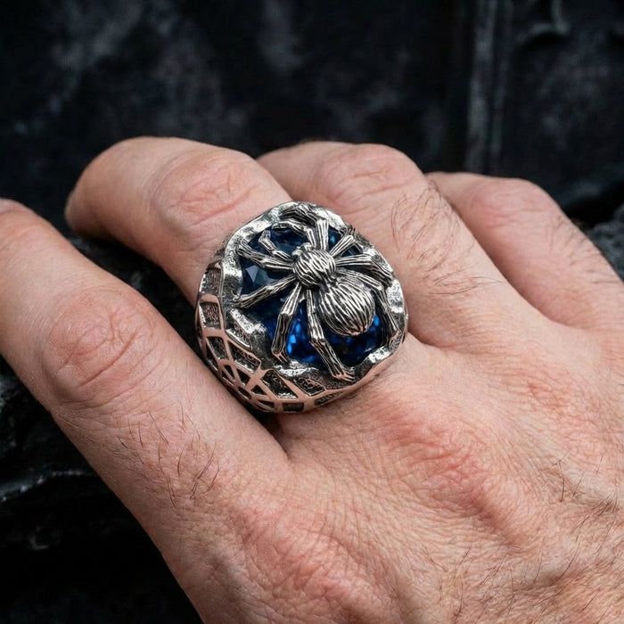 Men's gothic silver spider ring with large blue gemstone worn on hand, featuring detailed spider sculpture over deep blue stone with web-patterned band design on dark background