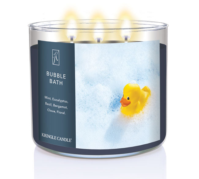 A three-wick lit candle in a clear glass jar with a calming bath-themed label. The candle’s wax is dark blue, and all three wicks are burning.