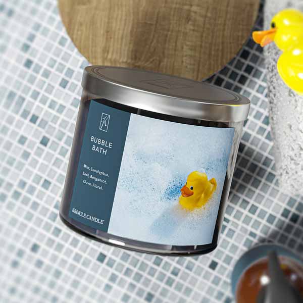 A three-wick candle in a clear glass jar with a silver lid and a calming bath-themed label. The label features a rubber duck floating in a sea of white bubbles. The candle’s wax is dark blue, and the wicks are unlit. The scent notes listed are mint, eucalyptus, basil, bergamot, clove, and floral.