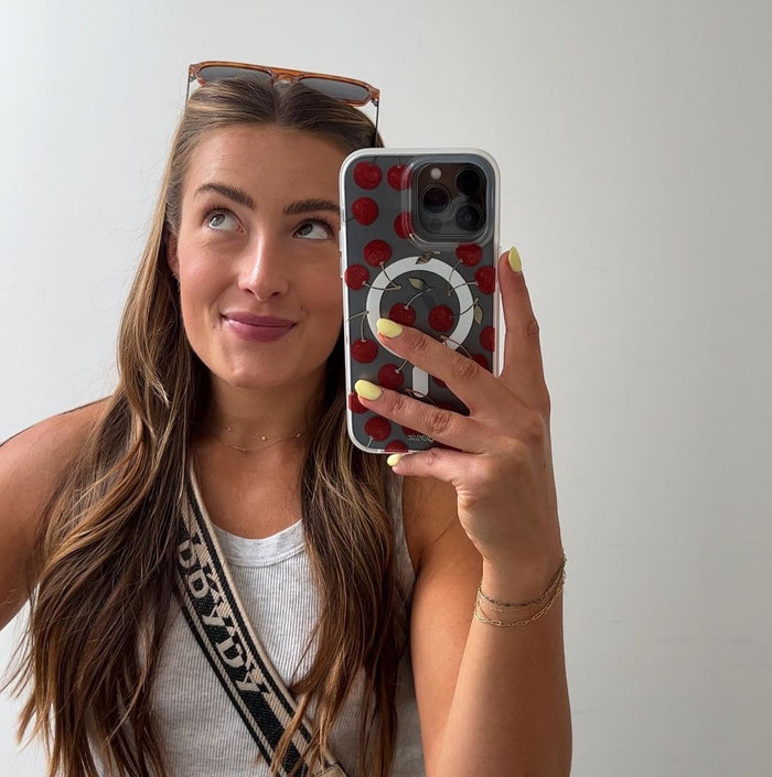 Front view of clear iPhone case with red cherry print and circular grip, held by a model in a mirror against a plain white wall