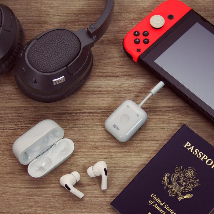 A flat lay photo featuring a passport, Matrix Cinema headphones, a handheld gaming console, a Connect Air wireless transmitter Bluetooth adapters, and earbuds on a wooden surface.