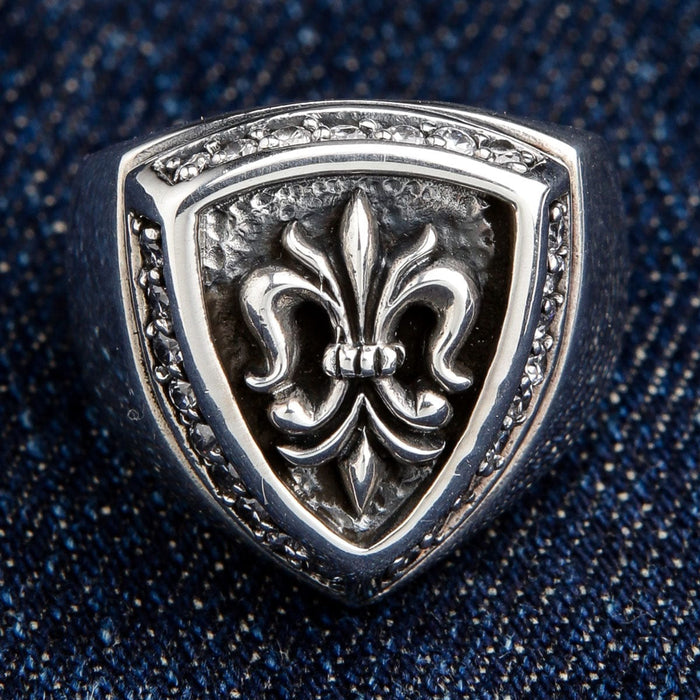 Front view of the men's fleur de lis shield ring resting on a blue denim texture.