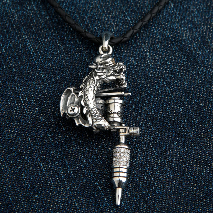 A sterling silver dragon tattoo machine pendant with a sparkling stone grip, shown on a black braided leather cord against a denim background.