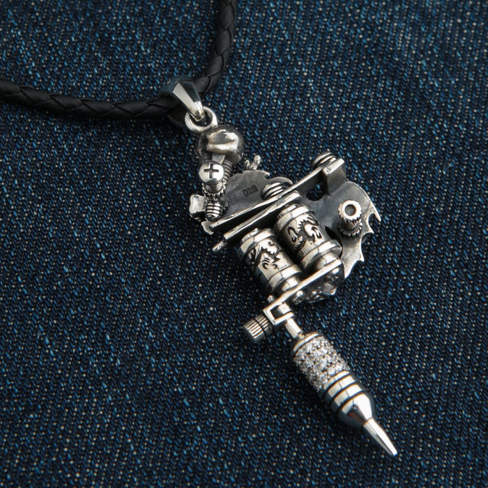 Tattoo-machine pendant angled on denim, engraved coils and a 925 stamp visible above the braided black cord.