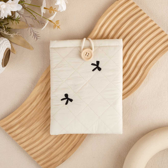 A white book sleeve with a embroidered bow design, closed with a button and elastic, displayed on a beige background with a wooden texture in the background.