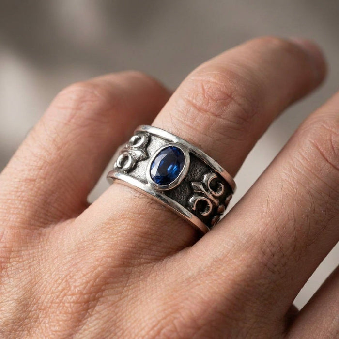 Modeled on a finger, the oxidized sterling silver ring displays its central blue stone and raised fleur-de-lis motifs.