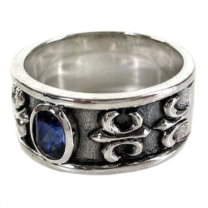 Angled perspective highlighting the dark oxidized band, raised silver motifs, and side-set blue cubic zirconia stone.