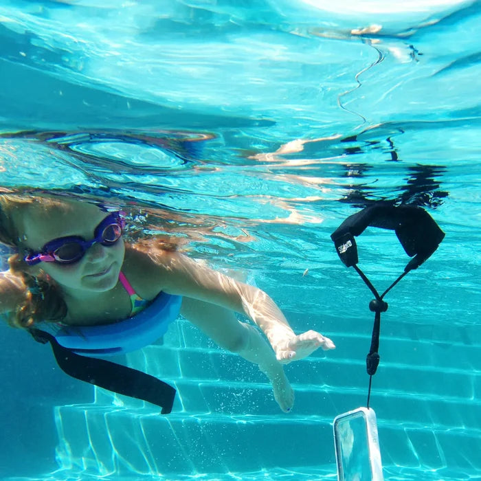 A young girl in purple goggles and a blue float swims underwater toward a Catalyst Case US Floating Lanyard - Stealth Black, which holds a phone case and floats nearby as sunlight reflects off the poolÃƒÂ¢Ã¢â€šÂ¬Ã¢â€žÂ¢s clear blue tiles.