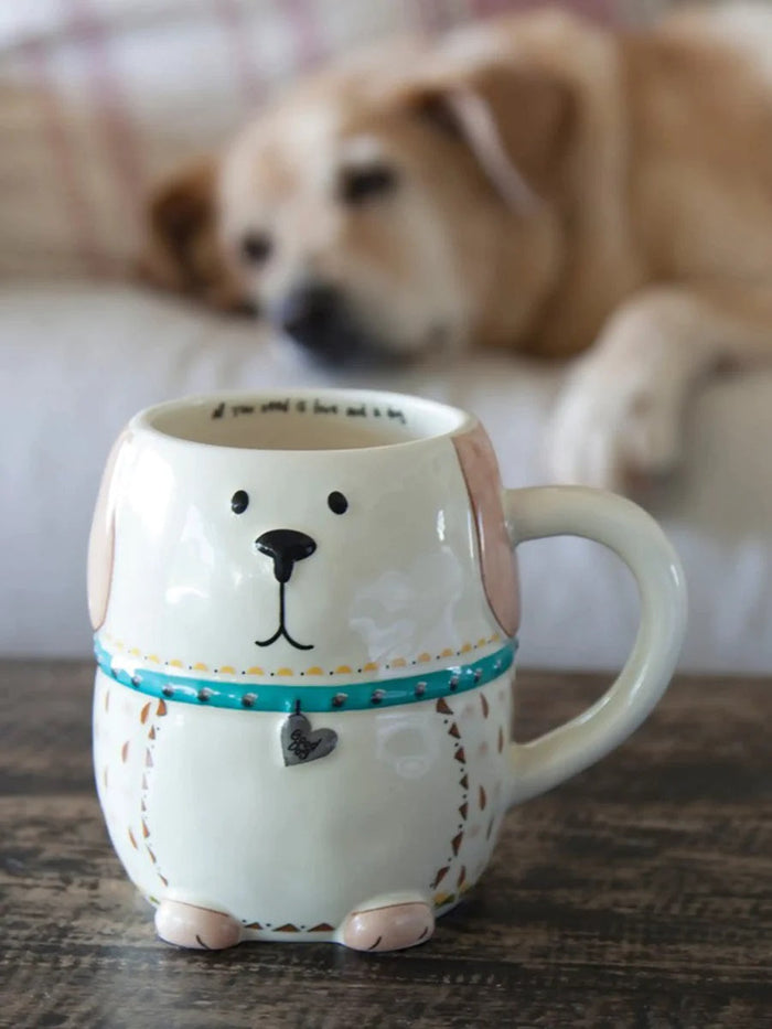 A ceramic mug designed to look like a dog, with a white body and a brown collar detail, positioned on a table with a sleeping dog in the background.