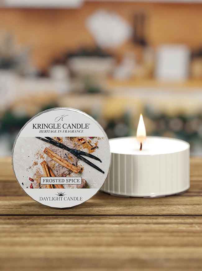 A lit white candle with a label reading "Kringle Candle, Frosted Spice" featuring cinnamon sticks and vanilla beans.