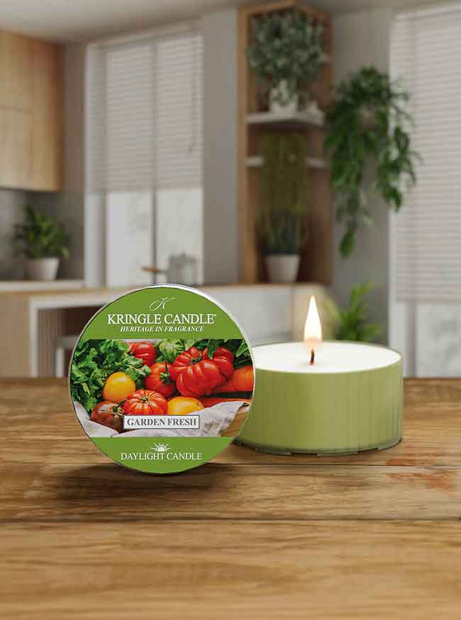 Green candle labeled "Garden Fresh" with tomatoes and greens, lit on a wooden table in a bright room with plants.