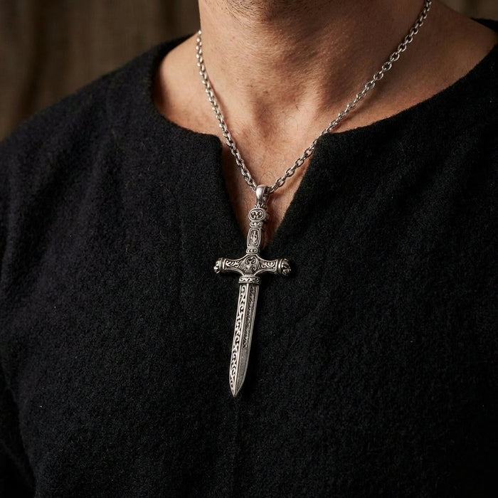 Sterling silver gothic dagger pendant with intricate medieval engravings on blade and crossguard, worn on thick chain necklace against black textured shirt on male model