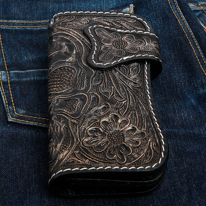 Skull and floral tooling worked into distressed dark leather, white stitching tracing the flap and metal loop.