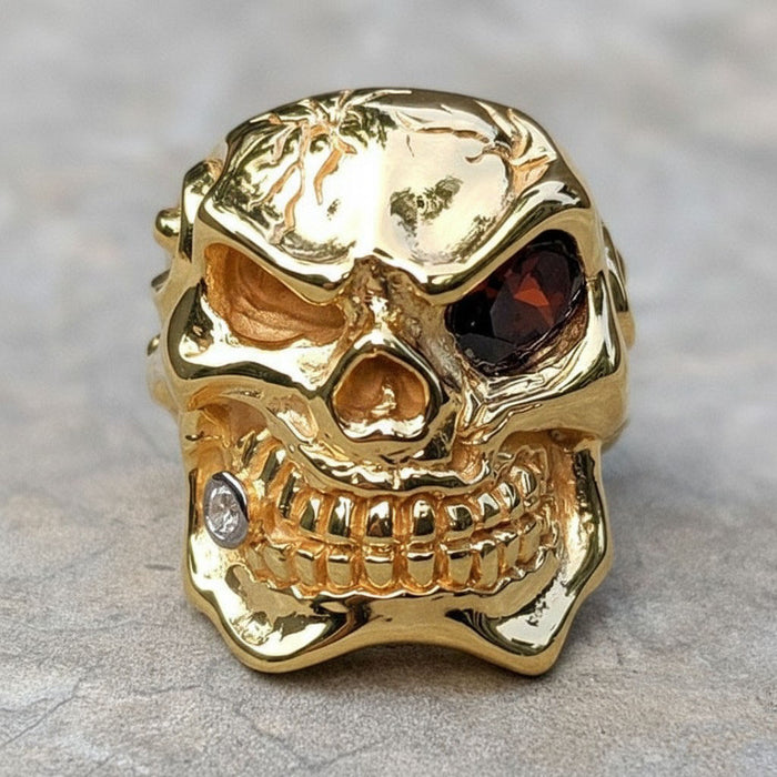 The front view of a heavy gold skull ring, showing a red gemstone eye, a cracked forehead, and a clear stone in one tooth.