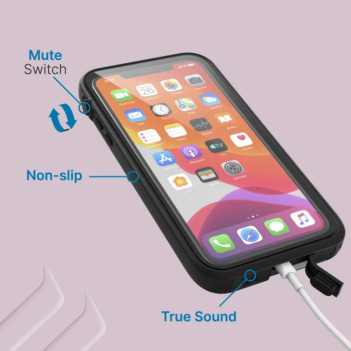 An iPhone 11 Pro Max in a black Catalyst waterproof case rests on a lavender surface. Labeled callouts highlight the mute switch, non-slip edges, and “True Sound” near the charging port with a cable plugged in; the screen shows colorful app icons.