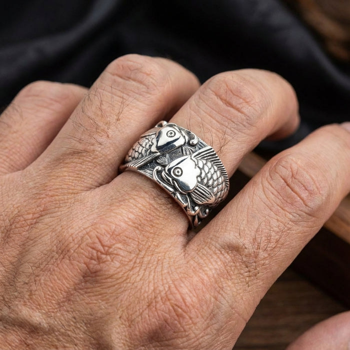 Close-up of a sterling silver Japanese Koi ring worn on a man's finger, featuring two detailed carp fish swimming around the wide band with oxidized scale textures.