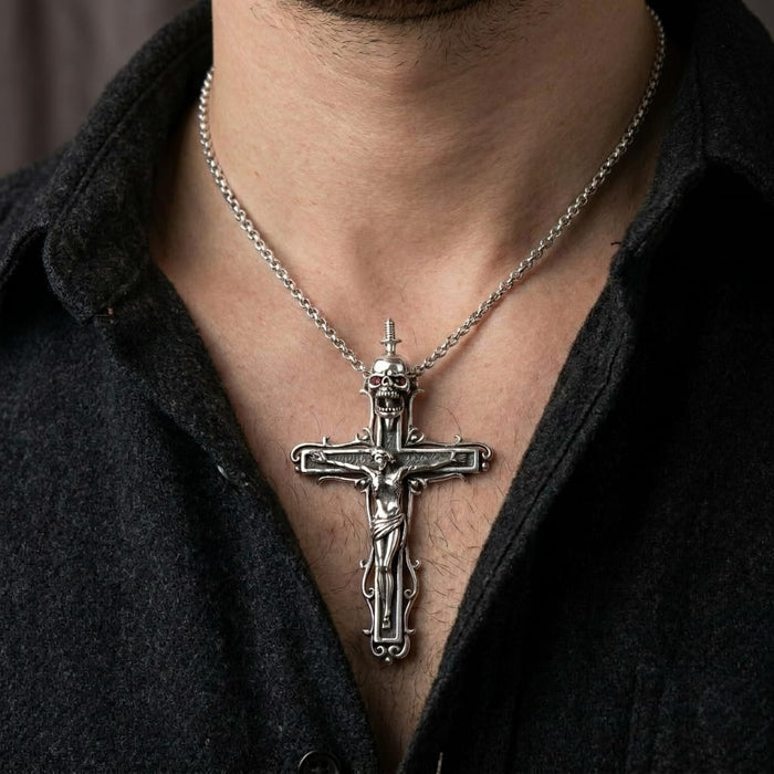 A close-up of a male model wearing an unbuttoned black shirt, showcasing a large, detailed sterling silver gothic pendant necklace featuring a crucifix topped with a skull that has red gemstone eyes.