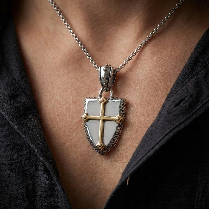 Silver knights shield pendant with gold cross detail worn on a silver chain around a man's neck, featuring medieval crusader-inspired design with textured edges and two-tone metalwork against an open dark shirt