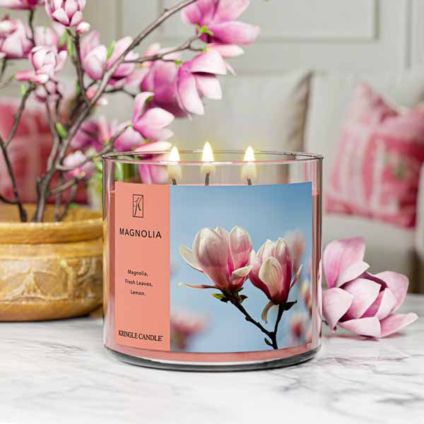 A three-wick lit candle in a clear glass jar with a soft floral-themed label. The candle’s wax is a warm peach shade, and all three wicks are burning.