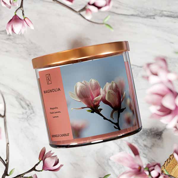 A three-wick candle in a clear glass jar with a bronze lid and a soft floral-themed label. The label features a close-up of pink magnolia blossoms against a bright blue sky. The candle’s wax is a warm peach shade, and the wicks are unlit. The scent notes listed are magnolia, fresh leaves, and lemon.