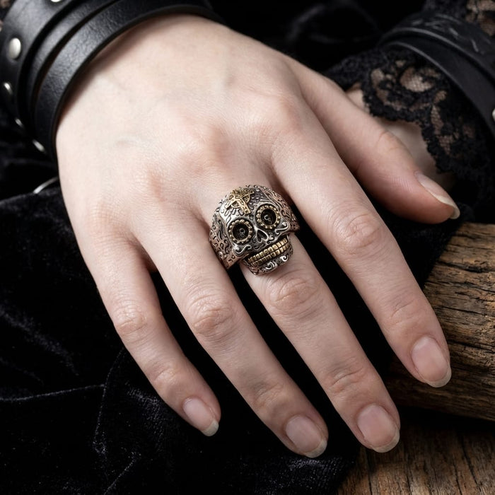 Close-up of a hand modeling an intricate silver and brass Mexican sugar skull ring, styled with black leather studded wristbands and dark velvet fabric for a gothic biker look.