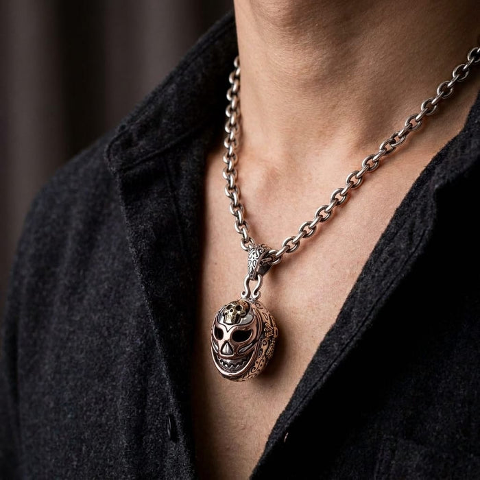 Man wearing a silver Mexican wrestler mask pendant on a thick chain over a dark shirt.