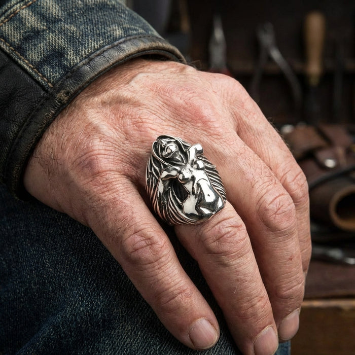 Large sterling silver nude lady ring with flowing hair design worn on a man's hand resting on denim jeans, showcasing detailed biker-style craftsmanship in a workshop setting