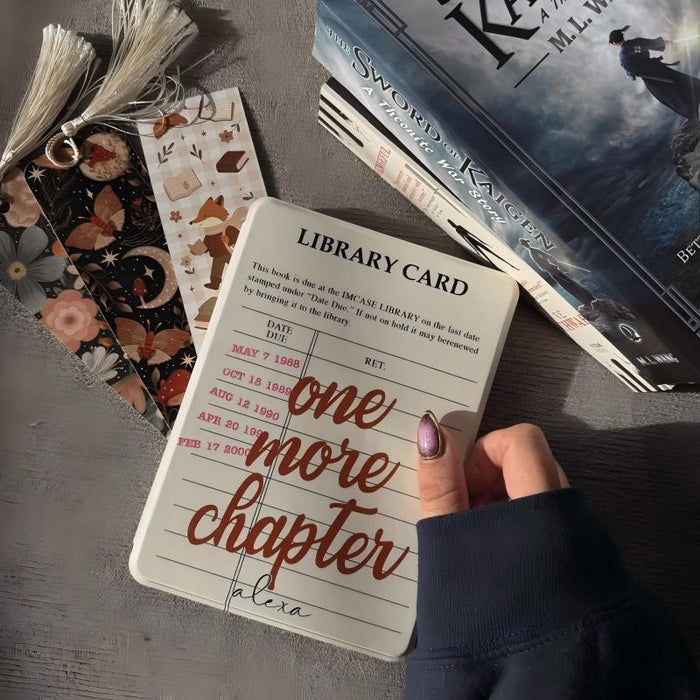 Library card with 'one more chapter' text held by a hand, surrounded by books and bookmarks.