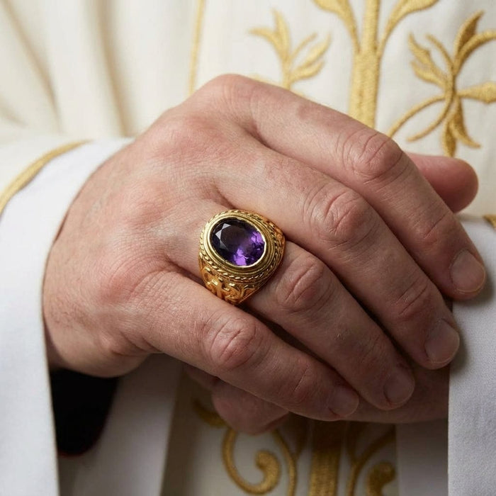Yellow gold bishop ring with large oval amethyst gemstone worn on hand over ornate gold-embroidered clergy vestments
