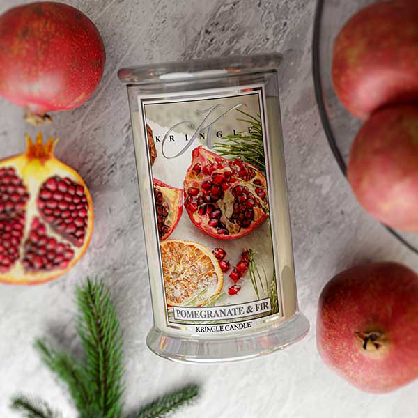 Kringle Candle with pomegranate and fir design surrounded by pomegranates and apples on a textured surface.