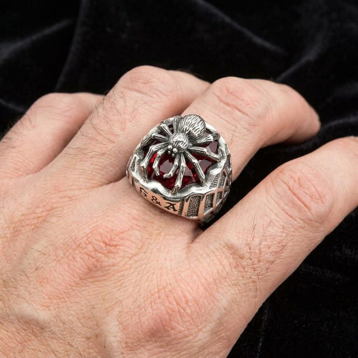 Man's hand wearing a silver gothic spider ring with a red stone against a dark background.