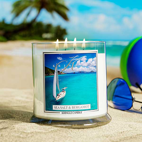 A lit Kringle Candle jar labeled “Sea Salt & Bergamot” sitting on sandy ground at the beach. The label shows a sailboat gliding across vibrant turquoise waters with mountains and clouds in the distance. A pair of blue-lensed sunglasses and a colorful beach ball rest nearby, with palm trees and ocean waves in the background, creating a tropical vibe.