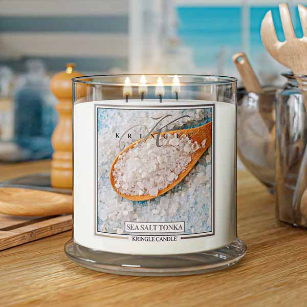 A lit Kringle Candle labeled “Sea Salt & Tonka” in a clear jar with four flames. The label features a close-up image of coarse sea salt in a wooden spoon resting on a bed of white and light blue salt crystals. The design evokes a fresh, clean, and coastal vibe against a plain white background.