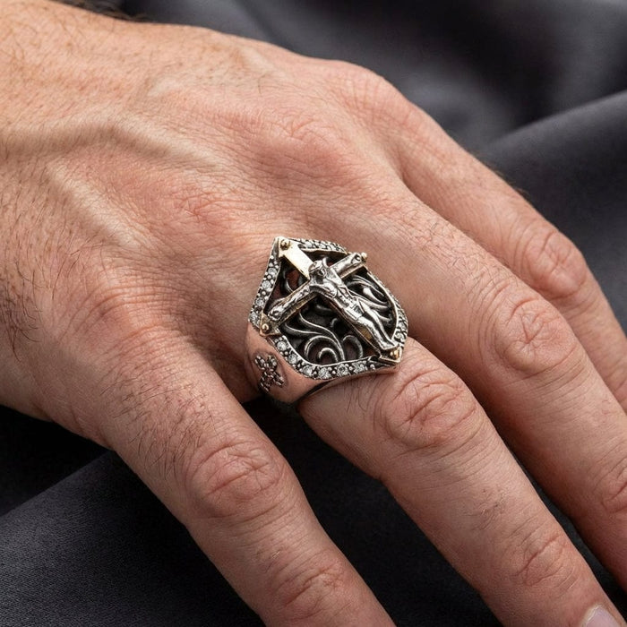 Sterling silver crucifix ring with shield-shaped design featuring a detailed Jesus on cross centerpiece, ornate scrollwork, and small diamond accents worn on a man's ring finger against dark fabric background