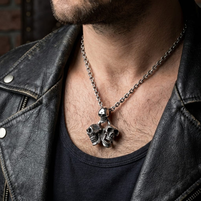 Close-up of a male model wearing a masculine silver triple skull pendant necklace on a chain, styled with a black shirt and a black leather biker jacket.