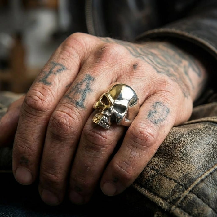 Close-up lifestyle shot of a rugged, tattooed hand wearing a large two-tone silver and brass skull biker ring, resting on a distressed brown leather jacket.
