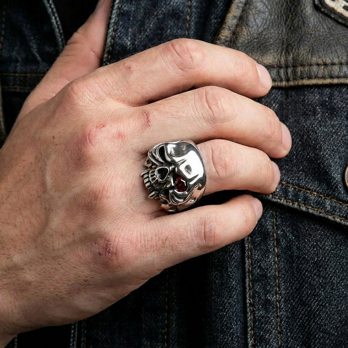 Silver vampire skull ring with red gemstone eye worn on a man's middle finger against a dark denim jacket, showcasing bold biker jewelry style