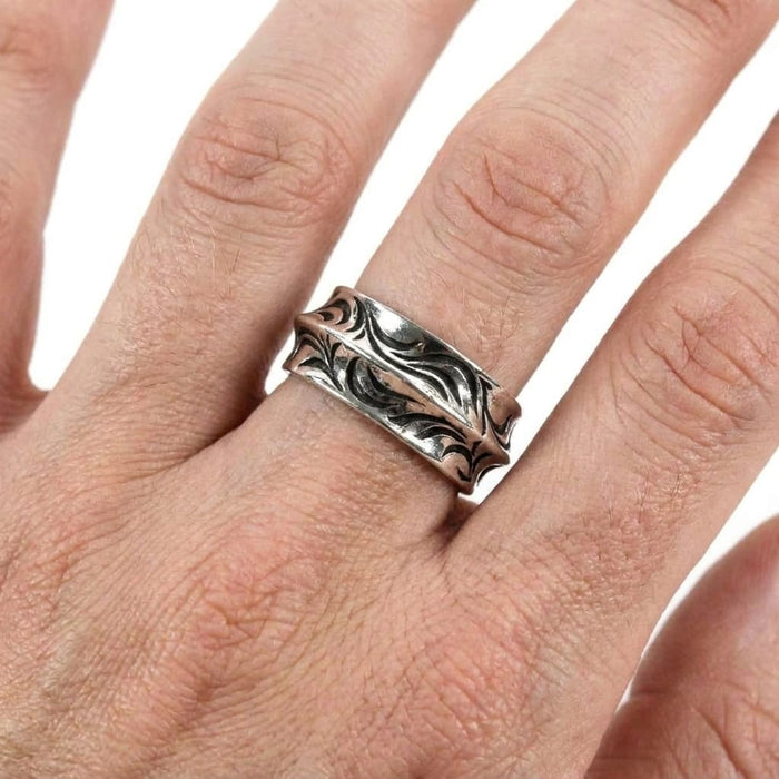 Close-up of a hand wearing a sterling silver band ring featuring an engraved, oxidized wave pattern against a white background.